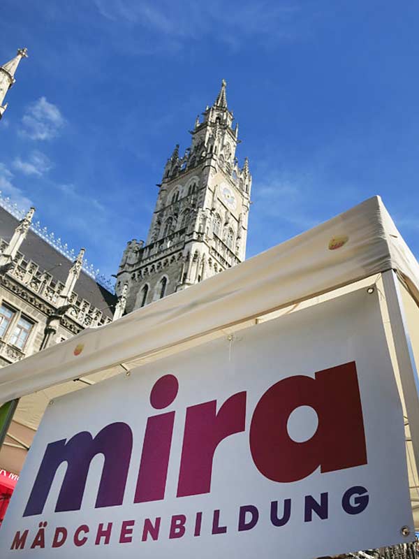 Links: Eine Fahne mit dem Schriftzug mira Mädchenbildung weht an einem weißen Infostand vor dem Münchner Rathaus. Rechts: 2 lilafarbene und ein schwarzer Luftballon mit der Aufschrift Internationaler Mädchentag vor blauem Himmel Links: Eine Fahne mit dem Schriftzug mira Mädchenbildung weht an einem weißen Infostand vor dem Münchner Rathaus. Rechts: 2 lilafarbene und ein schwarzer Luftballon mit der Aufschrift Internationaler Mädchentag vor blauem Himmel
