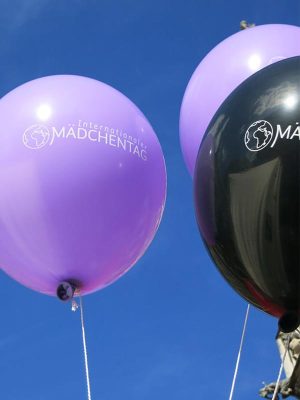 Zwei lilafarbene und ein schwarzer Luftballon mit der Aufschrift Internationaler Mädchentag vor blauem Himmel.