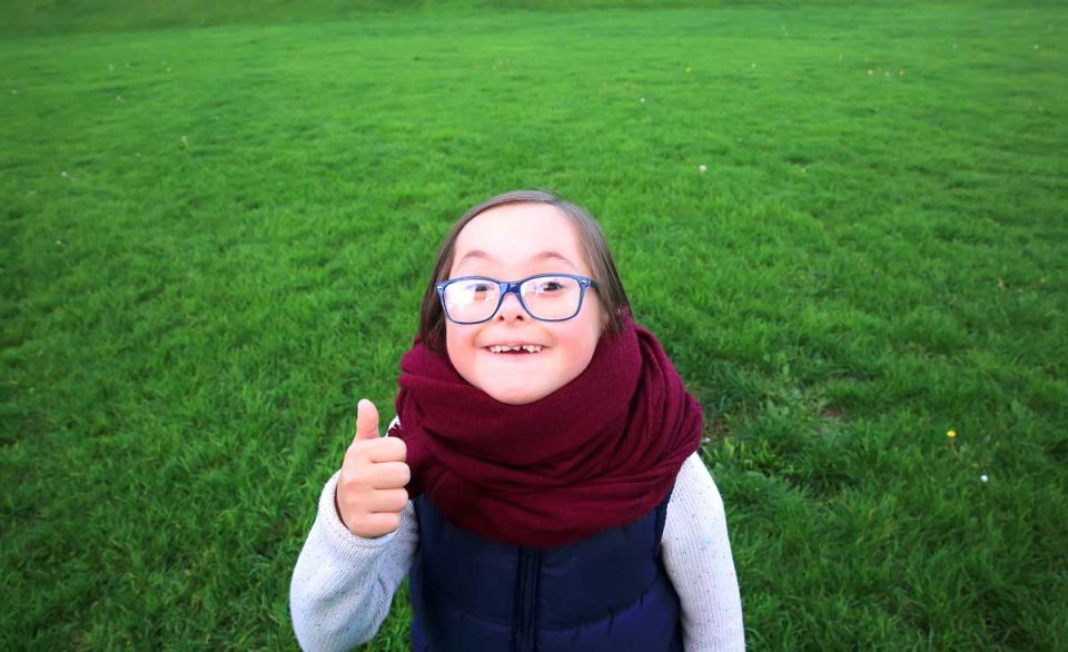 Ein Mädchen mit Down-Syndrom steht freudig auf einer grünen Wiese und zeigt die Geste: Daumen hoch!