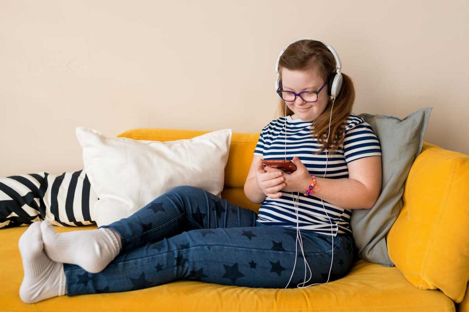 Eine junge Frau liegt entspannt auf dem Sofa, mit Kopfhörern auf, schaut auf den Display ihres Smartphones und lächelt dabei vergnügt.