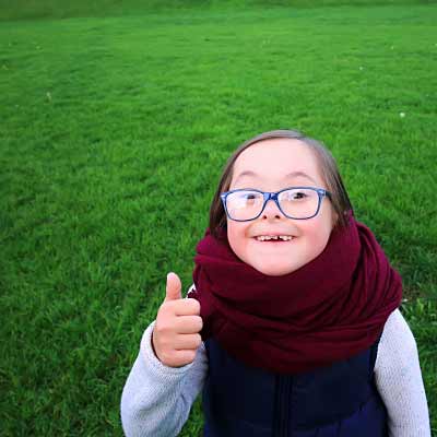 Ein Mädchen mit Down-Syndrom hebt den Daumen und lächelt in die Kamera.