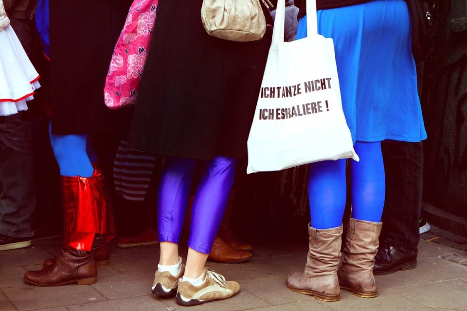 Junge Frauen in auffälligen, bunten Outfits stehen in einer Gruppe. Eine trägt eine Stofftasche mit Aufschrift: „ICH TANZE NICHT. ICH ESKALIERE!