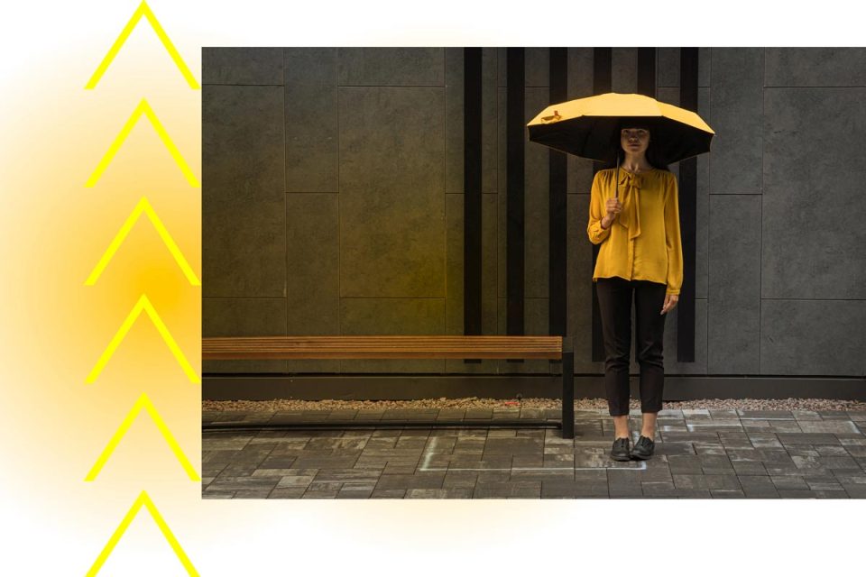 Eine junge Frau steht vor einer Gebäudewand unter einem gelben Regenschirm und schaut etwas betreten geradeaus in die Kamera.