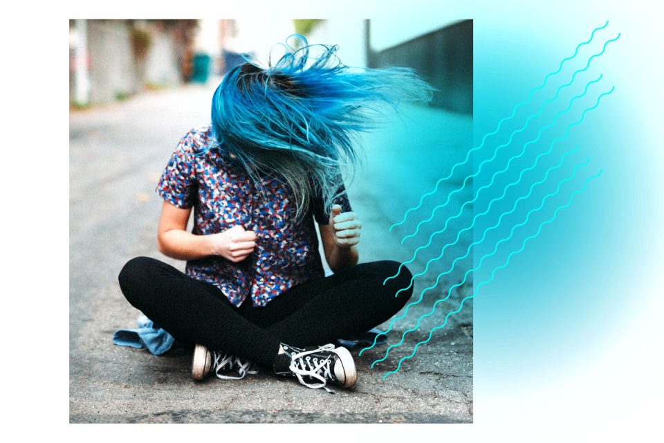 Eine junge Frau sitzt im Schneidersitz auf der Straße und schüttelt ihre langen blauen Haare.